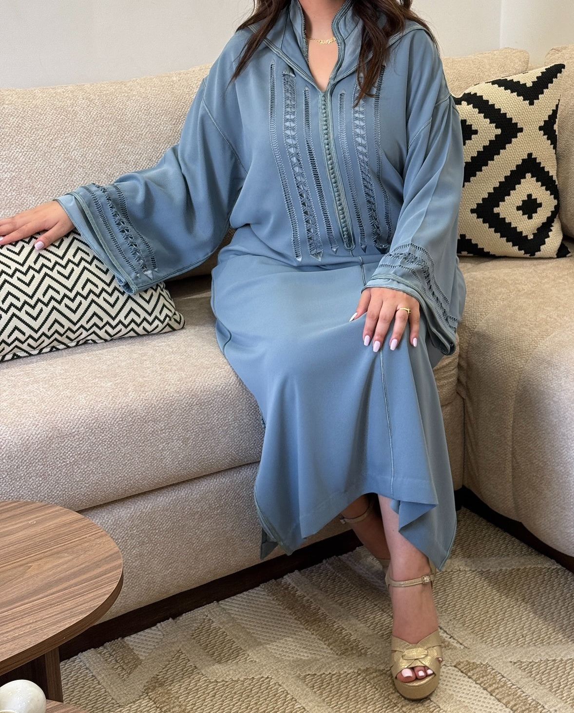 Woman in a blue djellaba sitting on a beige couch with patterned cushions.